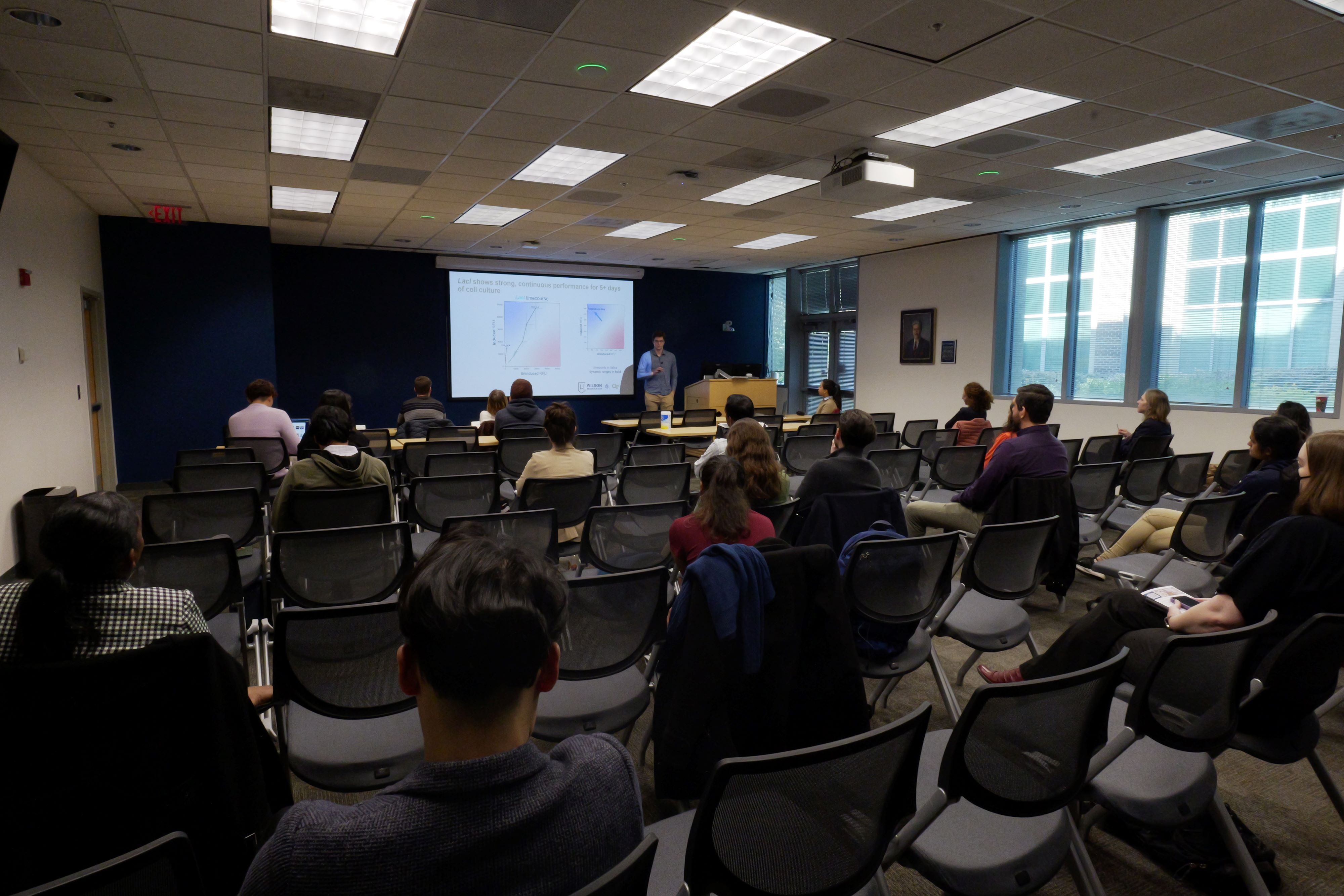 A man presenting at the front of a room with an audience.