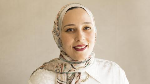A professional studio portrait of a woman smiling warmly, wearing a light-colored patterned hijab and a white collared blouse against a neutral, solid background.